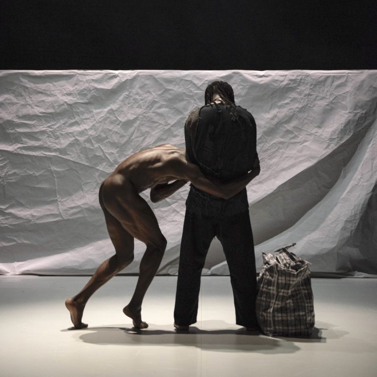 Two dancers in front of a white cloth background. One is naked with his back to the camera, the other has his head bowed down and is dressed in dark clothes