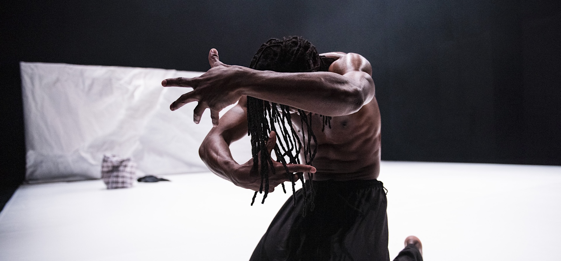 A black dancer with his arms partially covering his face poses before the camera on a white background