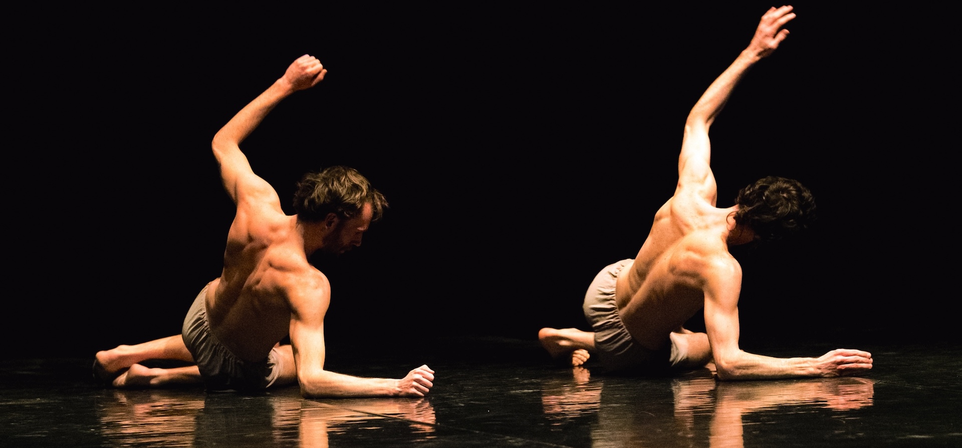 The backs of two dancers in a synchronised pose lying on the floor with one arm each up in the air