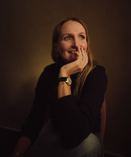 A headshot of Emma Miranda Moore with her chin resting on one hand. Background is dark with a gold glow.