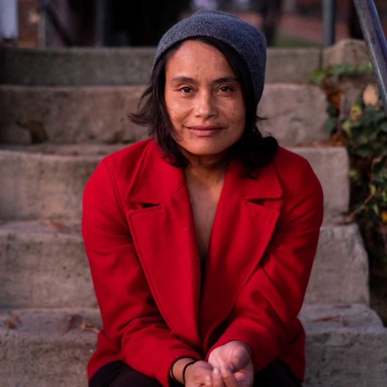 A headshot of Murielle Elizéon in a red coat sitting on concrete steps