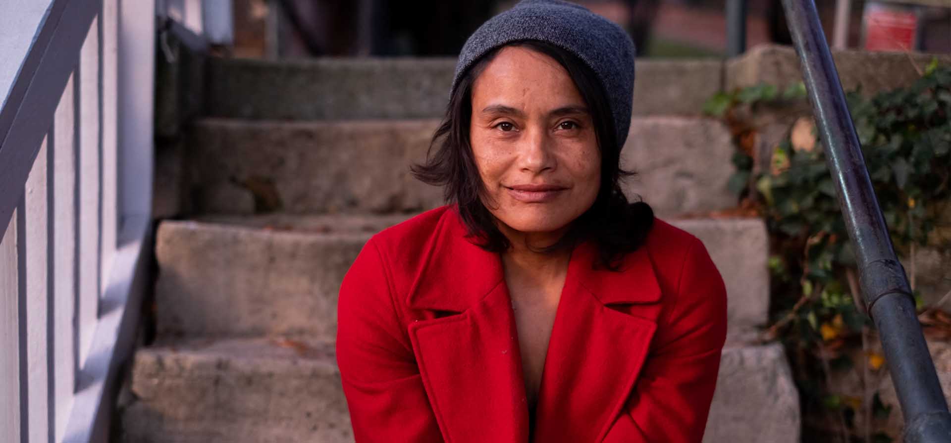 A headshot of Murielle Elizéon in a red coat sitting on concrete steps