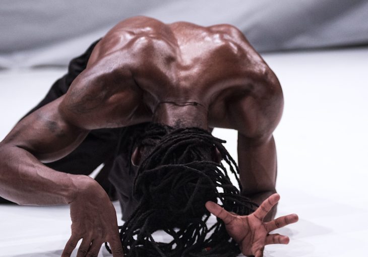 A black dancer with dreadlocks arches his back on the floor in the middle of dance routine