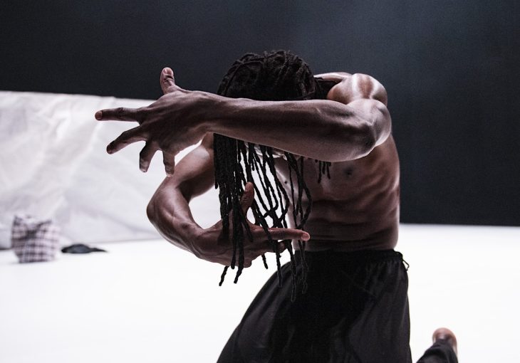 A black dancer with his arms partially covering his face poses before the camera on a white background