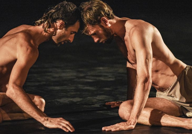 Two dancers sit on the floor in front a dark background touching their heads together