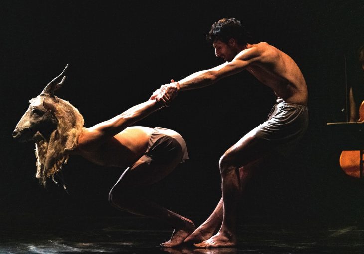 2 dancers in front of a dark background, one with an animal mask