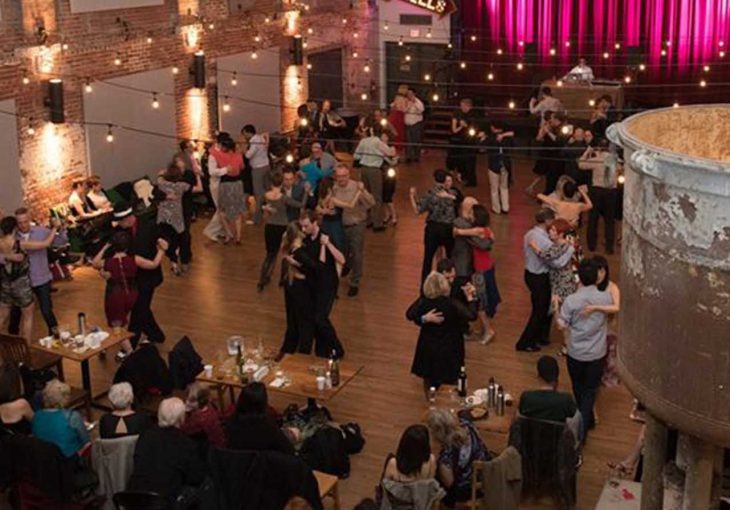 A landscape image of about 30 or 40 couples dancing the tango in a ballroom settings with soft lights